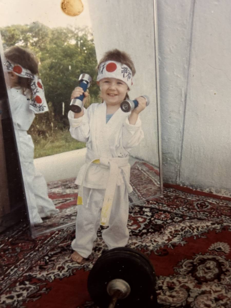 A young Rob in a gi and rising-sun headband, curling small dumbbells with a barbell on the floor.